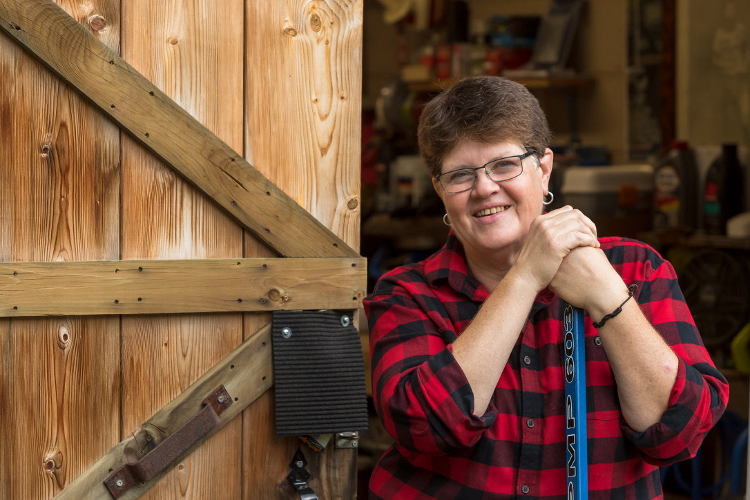 A cheerful woman in a red and black plaid shirt, leaning on a broom, standing next to a rustic wooden door. She has short brown hair, glasses, and a warm, approachable smile, with a cozy workshop visible in the background.