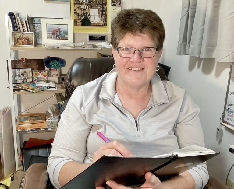 Tammy Watson, Canadian Handywoman, smiling while writing in a notebook at her desk with shelves of books and personal items behind her.